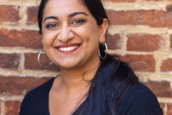 Sonia Kumar is a Middle Eastern woman with warm mediu brown skin tone. She is smiling at the camera and has long black hair pulled back in a low pony tail. She is wearing hoop earrings. She is standing in front of brick wall.