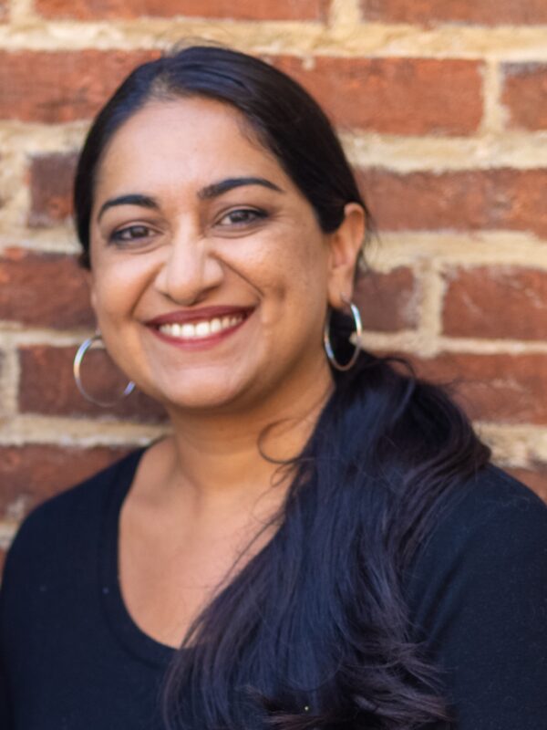 Sonia Kumar is a Middle Eastern woman with warm mediu brown skin tone. She is smiling at the camera and has long black hair pulled back in a low pony tail. She is wearing hoop earrings. She is standing in front of brick wall.