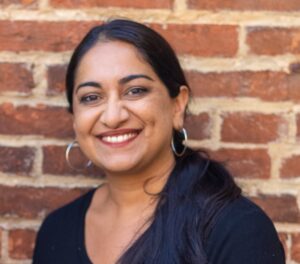 Sonia Kumar is a Middle Eastern woman with warm mediu brown skin tone. She is smiling at the camera and has long black hair pulled back in a low pony tail. She is wearing hoop earrings. She is standing in front of brick wall.