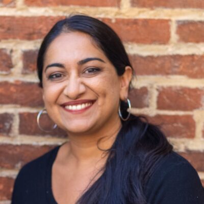 Sonia Kumar is a Middle Eastern woman with warm mediu brown skin tone. She is smiling at the camera and has long black hair pulled back in a low pony tail. She is wearing hoop earrings. She is standing in front of brick wall.