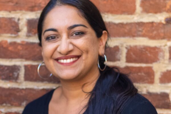 Sonia Kumar is a Middle Eastern woman with warm mediu brown skin tone. She is smiling at the camera and has long black hair pulled back in a low pony tail. She is wearing hoop earrings. She is standing in front of brick wall.