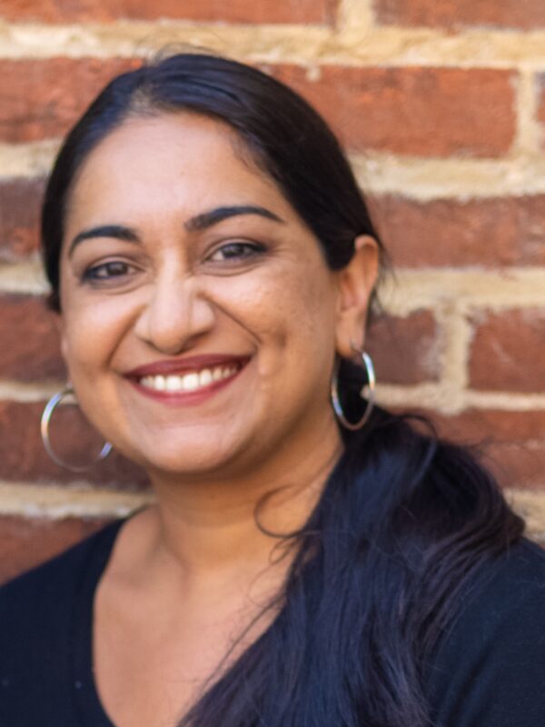 Sonia Kumar is a Middle Eastern woman with warm mediu brown skin tone. She is smiling at the camera and has long black hair pulled back in a low pony tail. She is wearing hoop earrings. She is standing in front of brick wall.