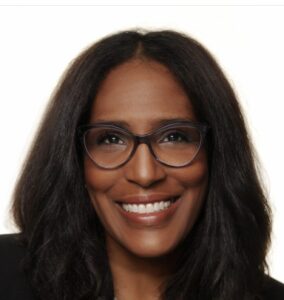 Shawn Essien is a Black woman who is smiling at the camera, has medium-length hair with light waves, rich warm Brown skin tone, is wearing glasses, a pearl necklace, and a black suit jacket.