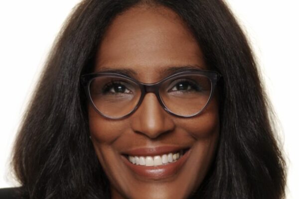Shawn Essien is a Black woman who is smiling at the camera, has medium-length hair with light waves, rich warm Brown skin tone, is wearing glasses, a pearl necklace, and a black suit jacket.