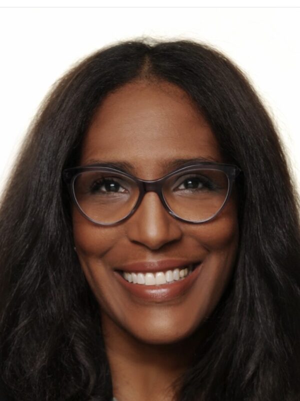 Shawn Essien is a Black woman who is smiling at the camera, has medium-length hair with light waves, rich warm Brown skin tone, is wearing glasses, a pearl necklace, and a black suit jacket.