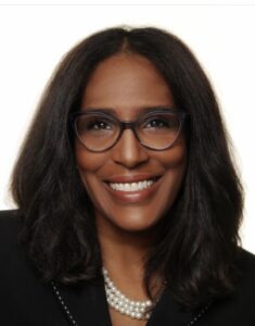 Shawn Essien is a Black woman who is smiling at the camera, has medium-length hair with light waves, rich warm Brown skin tone, is wearing glasses, a pearl necklace, and a black suit jacket.