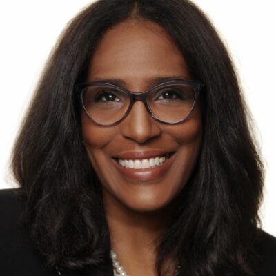 Shawn Essien is a Black woman who is smiling at the camera, has medium-length hair with light waves, rich warm Brown skin tone, is wearing glasses, a pearl necklace, and a black suit jacket.