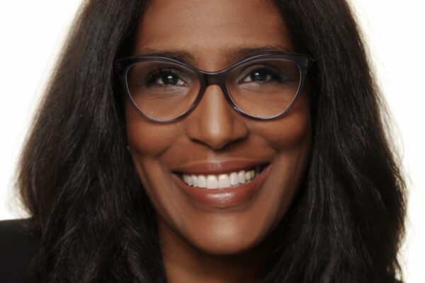 Shawn Essien is a Black woman who is smiling at the camera, has medium-length hair with light waves, rich warm Brown skin tone, is wearing glasses, a pearl necklace, and a black suit jacket.