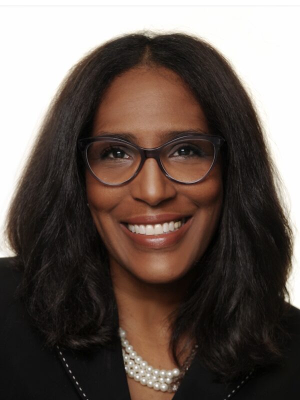 Shawn Essien is a Black woman who is smiling at the camera, has medium-length hair with light waves, rich warm Brown skin tone, is wearing glasses, a pearl necklace, and a black suit jacket.