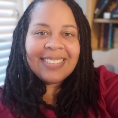 Erin Chapman is a woman with a latte skin tone, medium-length dark hair styled with locs, is smiling at the camera and wearing a red top.