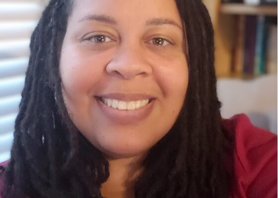 Erin Chapman is a woman with a latte skin tone, medium-length dark hair styled with locs, is smiling at the camera and wearing a red top.