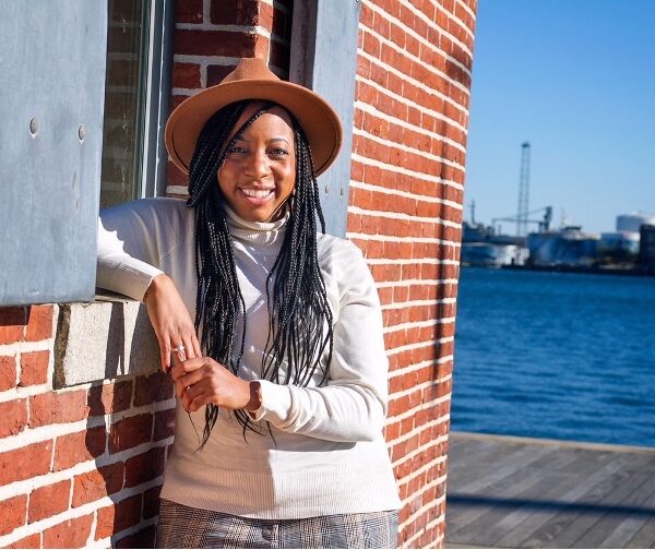 Oprah Keyes is a woman with chestnut skin tone, has long dark hair with small braided style. She is wearing a brimmed hat and white long sleep shirt. She is standing outside leaning on a brick wall with a dock and body of water in the background.