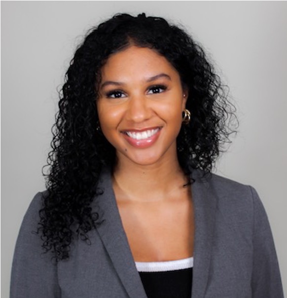 Yasmin Behram is a woman with a hazelnut skin tone, has medium-long curly dark hair, is wearing a gray blazer and black top with white piping. She is wearing rosy lipstick and is smiling at the camera. The background is a solid gray.