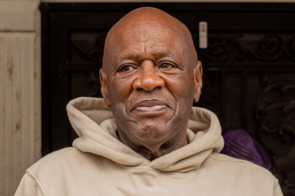 Bobby Stewart is a Black man with a beige hoodie, looking to the left, with a slight smile on his face. He is standing outside on a porch.