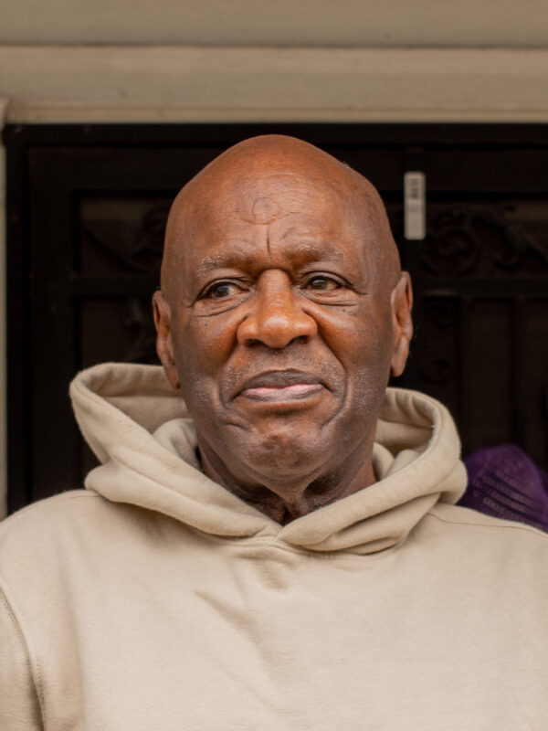 Bobby Stewart is a Black man with a beige hoodie, looking to the left, with a slight smile on his face. He is standing outside on a porch.