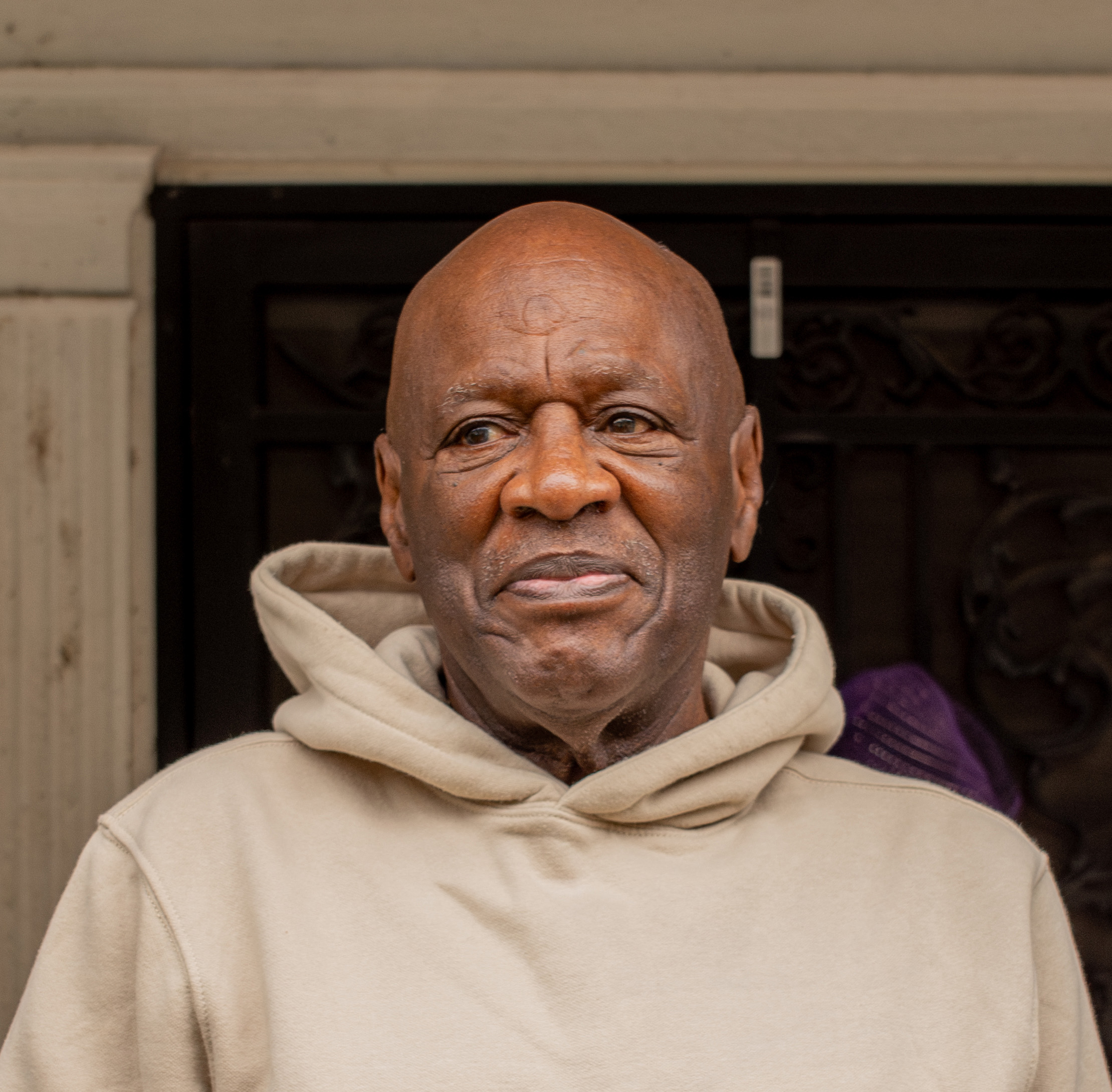 Bobby Stewart is a Black man with a beige hoodie, looking to the left, with a slight smile on his face. He is standing outside on a porch.