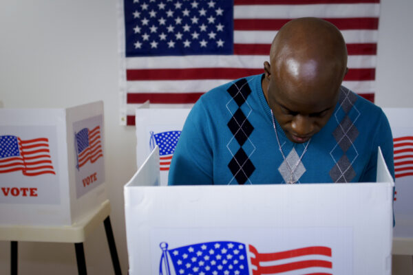 MS front view African American man in blue sweater and wearing a silver cross on a chain casting vote in booth at polling station. US flag on wall in background