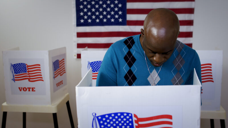 MS front view African American man in blue sweater and wearing a silver cross on a chain casting vote in booth at polling station. US flag on wall in background