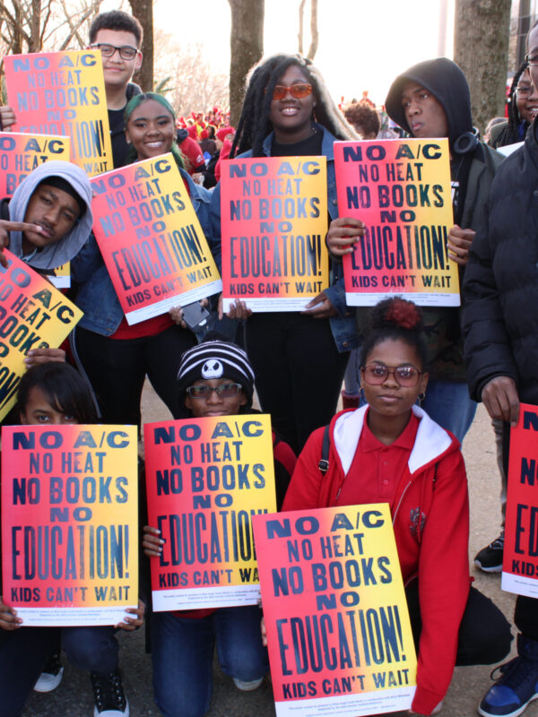 A group of students at Wide Angle Youth Media attend the March for Our Schools rally in Annapolis, Maryland. The students are holding posters they designed that are red and yellow gradient and say, "No A/C, no heat, no books, no education."