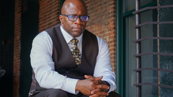 Anthony Muhammad is a Black man wearing glasses, shaved head, wearing a suit sitting on steps with a brick wall in the background.