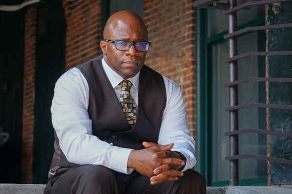 Anthony Muhammad is a Black man wearing glasses, shaved head, wearing a suit sitting on steps with a brick wall in the background.