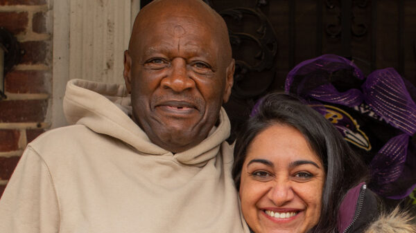 Bobby Stewart and Sonia Kumar are standing on a porch. Bobby has his arm around Sonia. Both are looking at the camera and smiling.