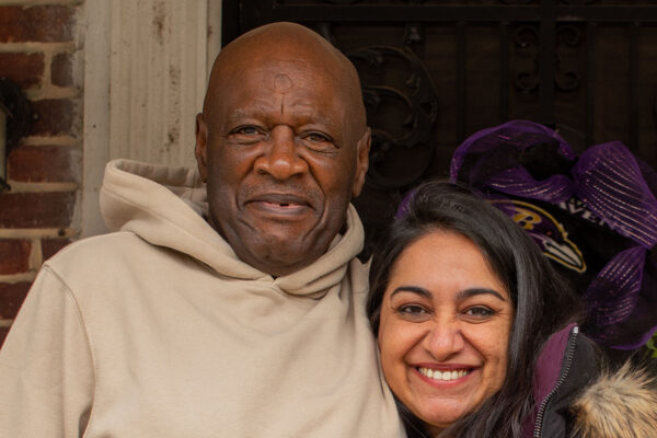 Bobby Stewart and Sonia Kumar are standing on a porch. Bobby has his arm around Sonia. Both are looking at the camera and smiling.