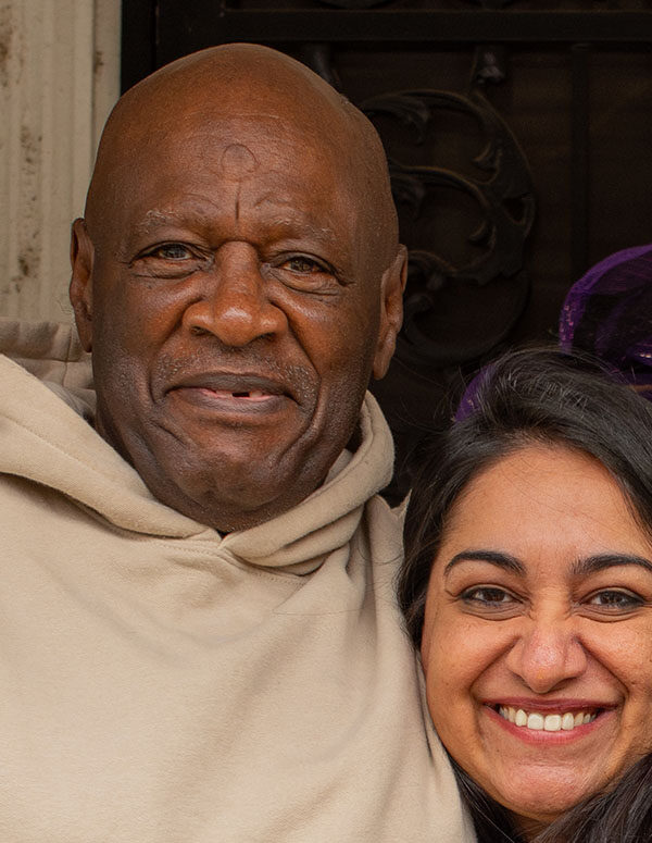 Bobby Stewart and Sonia Kumar are standing on a porch. Bobby has his arm around Sonia. Both are looking at the camera and smiling.