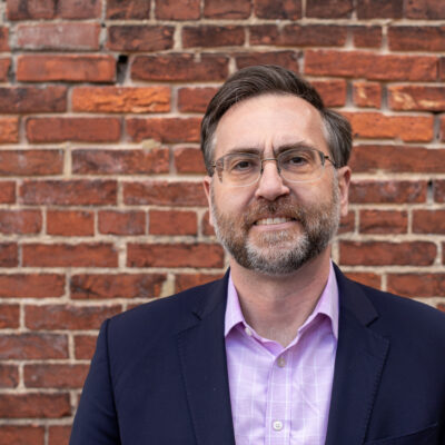 Corey Stottlemyer is standing in front of a brick wall. He is a white man with short brown and gray hair, wearing glasses and a suite.
