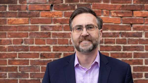 Corey Stottlemyer is standing in front of a brick wall. He is a white man with short brown and gray hair, wearing glasses and a suite.