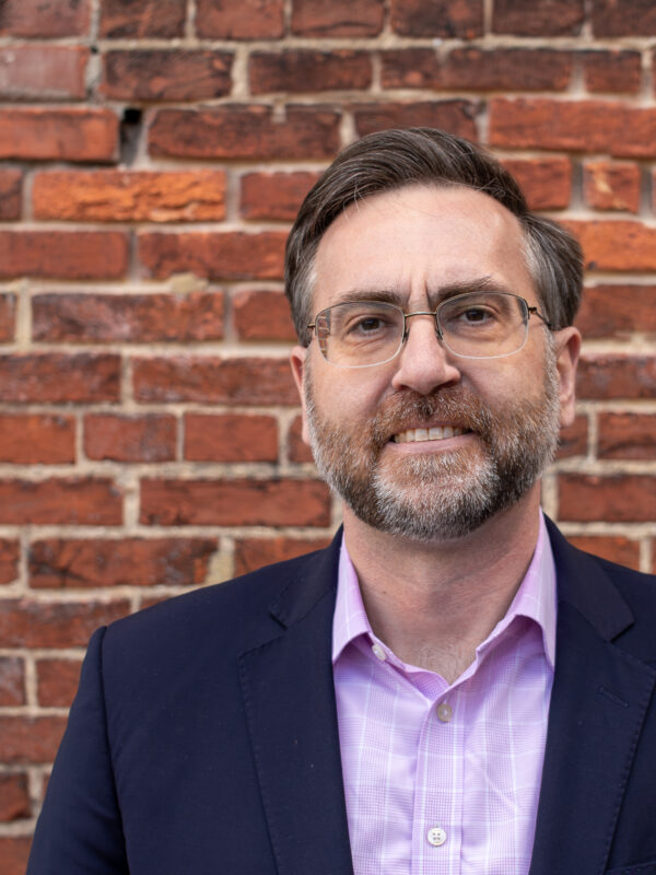 Corey Stottlemyer is standing in front of a brick wall. He is a white man with short brown and gray hair, wearing glasses and a suite.