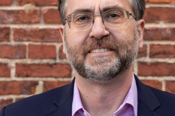 Corey Stottlemyer is standing in front of a brick wall. He is a white man with short brown and gray hair, wearing glasses and a suite.