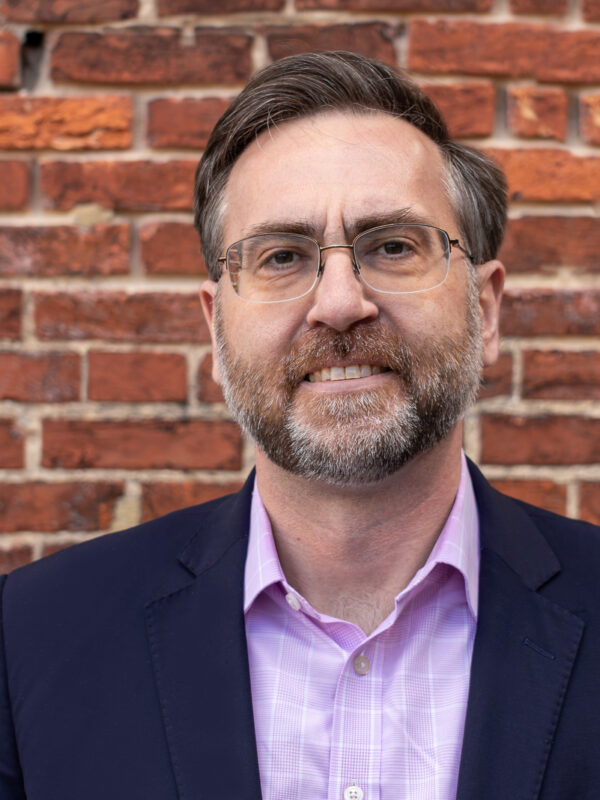 Corey Stottlemyer is standing in front of a brick wall. He is a white man with short brown and gray hair, wearing glasses and a suite.