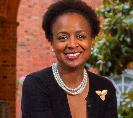 Kisha Petticolas is a Black woman with short natural hair and is smiling at the camera. She is wearing a pearl necklace and a dark cardigan. She is outside and has a brick wall and tree behind her.