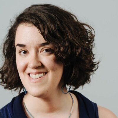 Sarah Dwyer is a white woman with chin-length dark wavy hair. She is smiling at the camera and is wearing a dark sleeveless top. The background is a gray wall.