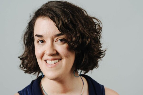 Sarah Dwyer is a white woman with chin-length dark wavy hair. She is smiling at the camera and is wearing a dark sleeveless top. The background is a gray wall.