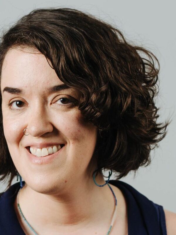 Sarah Dwyer is a white woman with chin-length dark wavy hair. She is smiling at the camera and is wearing a dark sleeveless top. The background is a gray wall.