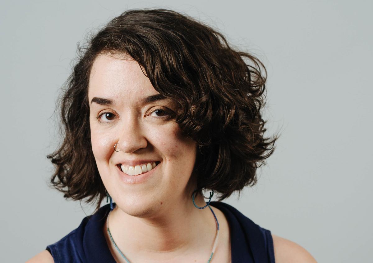 Sarah Dwyer is a white woman with chin-length dark wavy hair. She is smiling at the camera and is wearing a dark sleeveless top. The background is a gray wall.