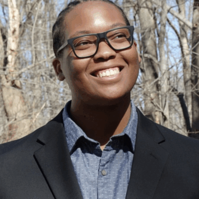Carlos Childs is looping to the side and up and is smiling. He is a Black man and is earing dark0framed glasses. He is outside in front of trees, is wearing a black suit jacket over a blue button-up and collared shirt.