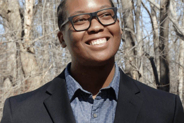 Carlos Childs is looping to the side and up and is smiling. He is a Black man and is earing dark0framed glasses. He is outside in front of trees, is wearing a black suit jacket over a blue button-up and collared shirt.