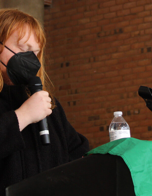 Chessie Jackson has shoulder-length red hair, is wearing a mask, and is speaking into a microphone. There is a Palestinian flag draped next to her.