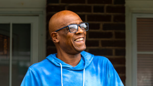 Wayne Brewton-Bey is a Black man with warm medium-brown skin tone, has shaved head, is wearing glasses, and is smiling. He is standing outside on a front porch.