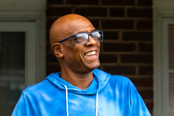 Wayne Brewton-Bey is a Black man with warm medium-brown skin tone, has shaved head, is wearing glasses, and is smiling. He is standing outside on a front porch.