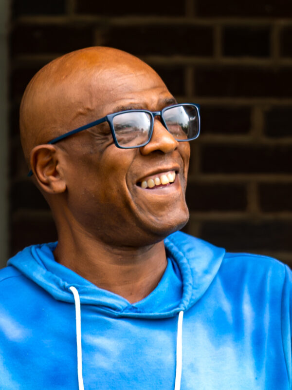 Wayne Brewton-Bey is a Black man with warm medium-brown skin tone, has shaved head, is wearing glasses, and is smiling. He is standing outside on a front porch.