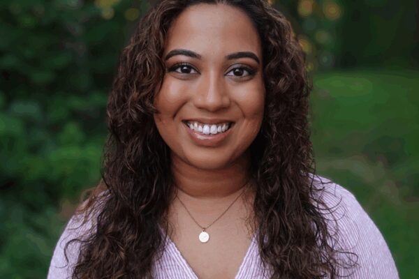 Fathima Rifkey is standing outside with trees and grassy field in the background. She has warm medium-brown skin, long curly hair, is smiling at the camera, and is wearing a pink v-neck shirt.
