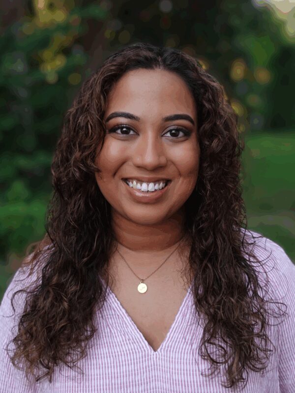 Fathima Rifkey is standing outside with trees and grassy field in the background. She has warm medium-brown skin, long curly hair, is smiling at the camera, and is wearing a pink v-neck shirt.
