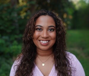 Fathima Rifkey is standing outside with trees and grassy field in the background. She has warm medium-brown skin, long curly hair, is smiling at the camera, and is wearing a pink v-neck shirt.