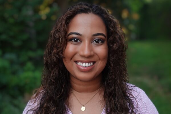 Fathima Rifkey is standing outside with trees and grassy field in the background. She has warm medium-brown skin, long curly hair, is smiling at the camera, and is wearing a pink v-neck shirt.