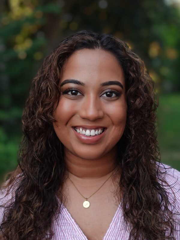 Fathima Rifkey is standing outside with trees and grassy field in the background. She has warm medium-brown skin, long curly hair, is smiling at the camera, and is wearing a pink v-neck shirt.