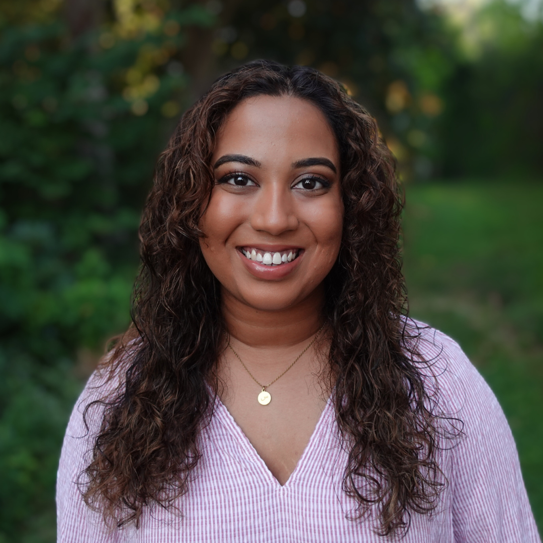 Fathima Rifkey is standing outside with trees and grassy field in the background. She has warm medium-brown skin, long curly hair, is smiling at the camera, and is wearing a pink v-neck shirt.